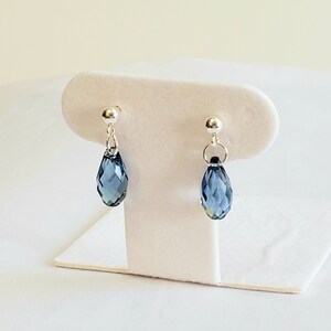 May include: A pair of silver-toned drop earrings. Each earring features a small, round stud and a teardrop-shaped, faceted blue crystal pendant. The earrings are displayed on a white jewelry stand against a soft, neutral background.