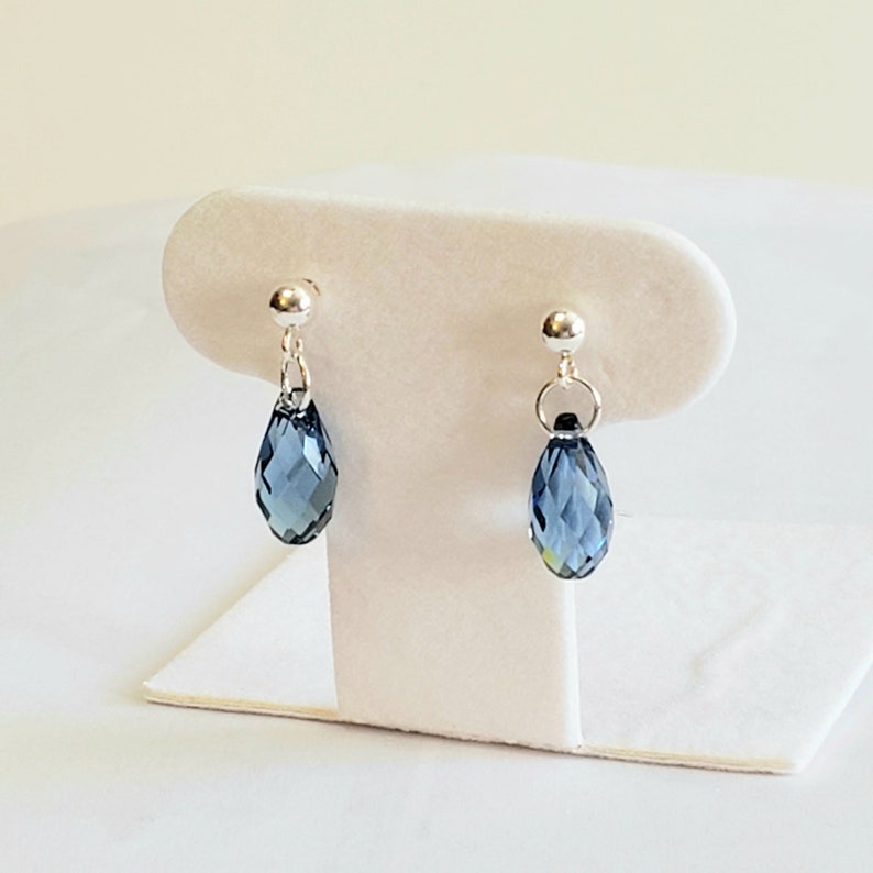 May include: A pair of silver-toned drop earrings. Each earring features a small, round stud and a teardrop-shaped, faceted blue crystal pendant. The earrings are displayed on a white jewelry stand against a soft, neutral background.