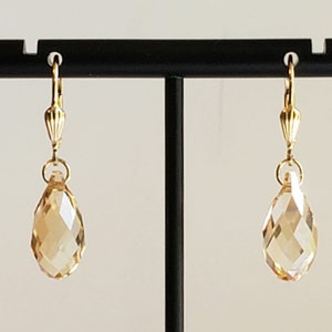 May include: A pair of gold-toned leverback earrings featuring faceted, teardrop-shaped champagne-coloured crystals. The crystals are suspended from a ribbed gold-toned metal finding, creating a clear, reflective, and symmetrical drop earring design.