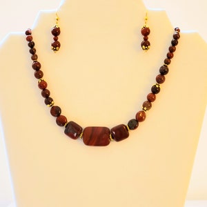 May include: A red and brown gemstone necklace and earrings set. The necklace features a large rectangular red gemstone in the center, surrounded by smaller round red and brown gemstones. The earrings are simple and feature a single round red gemstone.