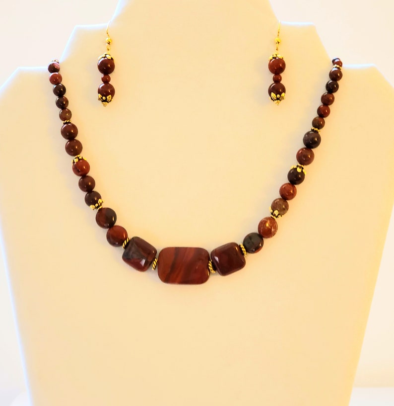 May include: A red and brown gemstone necklace and earrings set. The necklace features a large rectangular red gemstone in the center, surrounded by smaller round red and brown gemstones. The earrings are simple and feature a single round red gemstone.