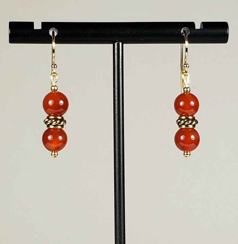 May include: A pair of drop earrings featuring two smooth, round, deep orange-red carnelian beads separated by a small, textured gold-tone metal spacer. The beads are suspended from gold-tone metal ear wires, creating a vertical, symmetrical design.