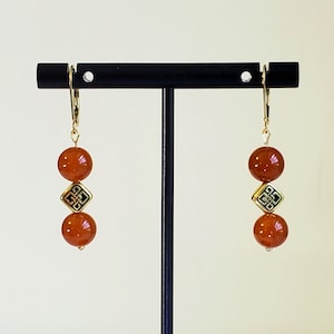 May include: A pair of drop earrings featuring two round, polished, burnt-orange beads separated by a small, square, gold-toned metal charm with a black, intricate, geometric knot pattern. The earrings hang from gold-toned lever-back closures.