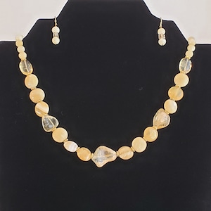 May include: A gold necklace with a light yellow and white stone bead design. The necklace is on a black mannequin bust. The necklace is paired with matching earrings.
