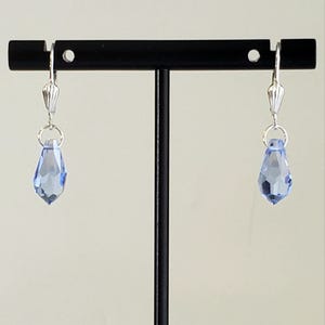 May include: A pair of light blue teardrop earrings with silver-colored hardware. The earrings are displayed on a black stand against a neutral background. The faceted crystal drops catch the light, showcasing their color and shape.