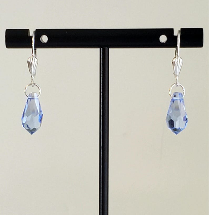 May include: A pair of light blue teardrop earrings with silver-colored hardware. The earrings are displayed on a black stand against a neutral background. The faceted crystal drops catch the light, showcasing their color and shape.