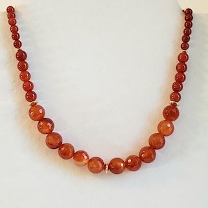 May include: A necklace featuring a strand of round, deep orange carnelian beads. The beads transition from smaller, smooth spheres near the ends to larger, faceted spheres toward the center, accented by small, polished gold-tone metal spacers.