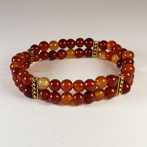 May include: A double-strand bracelet featuring polished, round, reddish-orange carnelian beads. The two rows of beads are connected by two rectangular, gold-toned metal spacers with a textured, beaded design on their vertical edges.