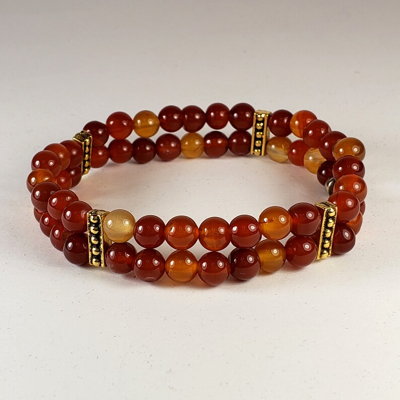 May include: A double-strand bracelet featuring polished, round, reddish-orange carnelian beads. The two rows of beads are connected by two rectangular, gold-toned metal spacers with a textured, beaded design on their vertical edges.