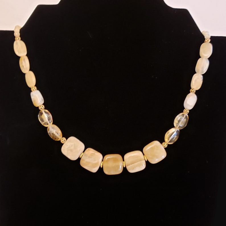 May include: A necklace featuring a variety of light yellow and clear beads. The necklace has a gold-colored chain and clasp. The beads are a mix of square and oval shapes. The necklace is displayed on a black background.