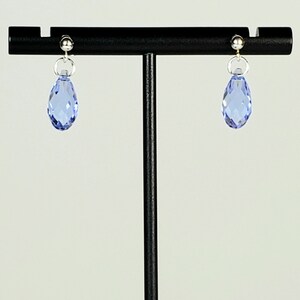 May include: A pair of light blue teardrop earrings. Each earring features a faceted, light blue crystal pendant suspended from a silver-colored post and ring. The earrings are displayed on a black stand against a white background.