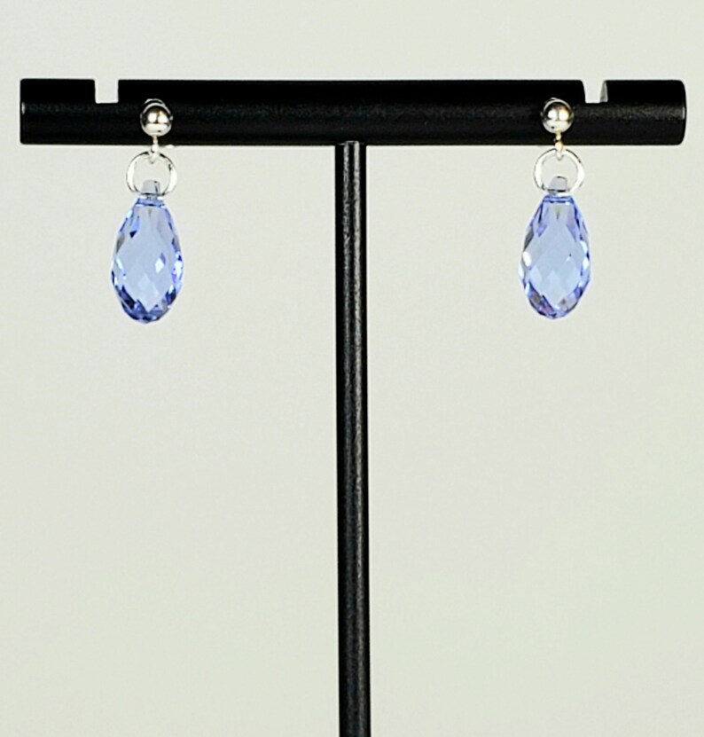 May include: A pair of light blue teardrop earrings. Each earring features a faceted, light blue crystal pendant suspended from a silver-colored post and ring. The earrings are displayed on a black stand against a white background.