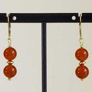 May include: A pair of drop earrings featuring two round, translucent orange beads separated by a small gold-toned spacer bead. The beads are suspended from gold-toned leverback ear wires, creating a simple, symmetrical design with warm tones.