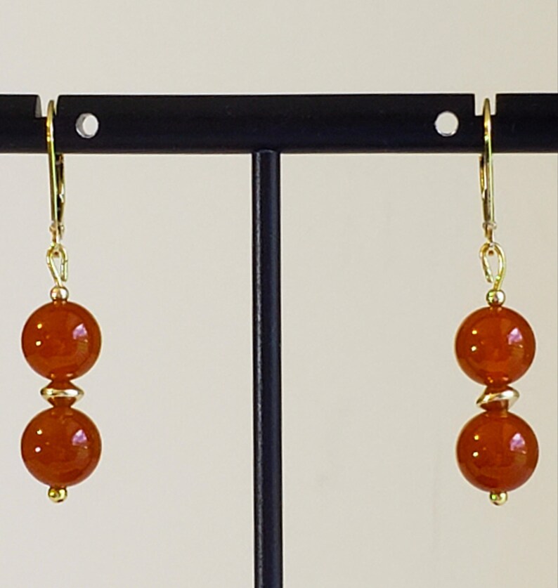 May include: A pair of drop earrings featuring two round, translucent orange beads separated by a small gold-toned spacer bead. The beads are suspended from gold-toned leverback ear wires, creating a simple, symmetrical design with warm tones.