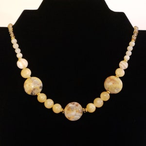 May include: A beaded necklace featuring large, round, speckled beige stones interspersed with smaller, round, yellow beads. The necklace has gold-colored accents and is displayed against a black background.