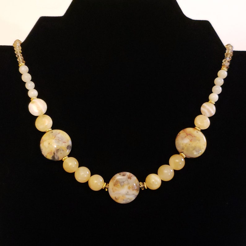 May include: A beaded necklace featuring large, round, speckled beige stones interspersed with smaller, round, yellow beads. The necklace has gold-colored accents and is displayed against a black background.
