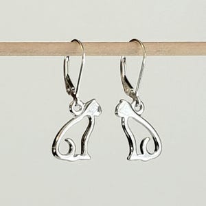 May include: A pair of silver-colored cat-shaped earrings. Each earring features a lever-back closure and a cutout design of a sitting cat. The earrings are displayed on a wooden dowel against a neutral background.