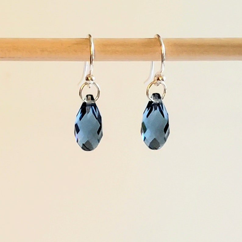 May include: A pair of teardrop-shaped earrings with faceted, dark blue crystals. The crystals hang from silver-colored ear wires. The earrings are displayed against a light background, highlighting their color and shape.