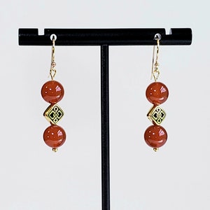 May include: A pair of drop earrings featuring two smooth, round, reddish-brown beads separated by a single gold-toned square bead with a black etched knot pattern. The earrings hang from gold-toned metal hooks and are displayed on a black stand.