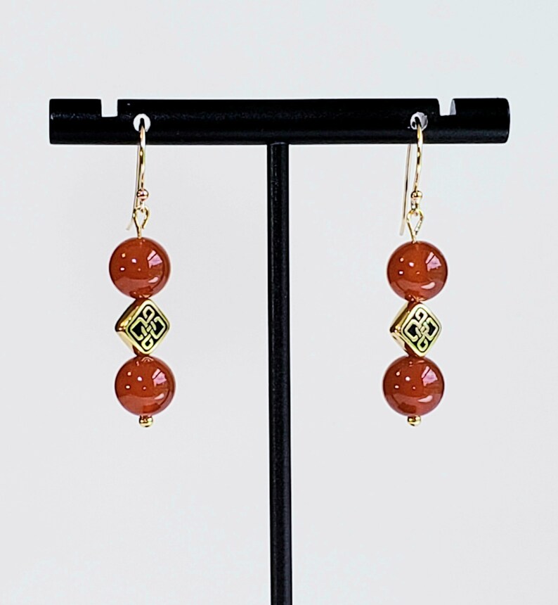 May include: A pair of drop earrings featuring two smooth, round, reddish-brown beads separated by a single gold-toned square bead with a black etched knot pattern. The earrings hang from gold-toned metal hooks and are displayed on a black stand.