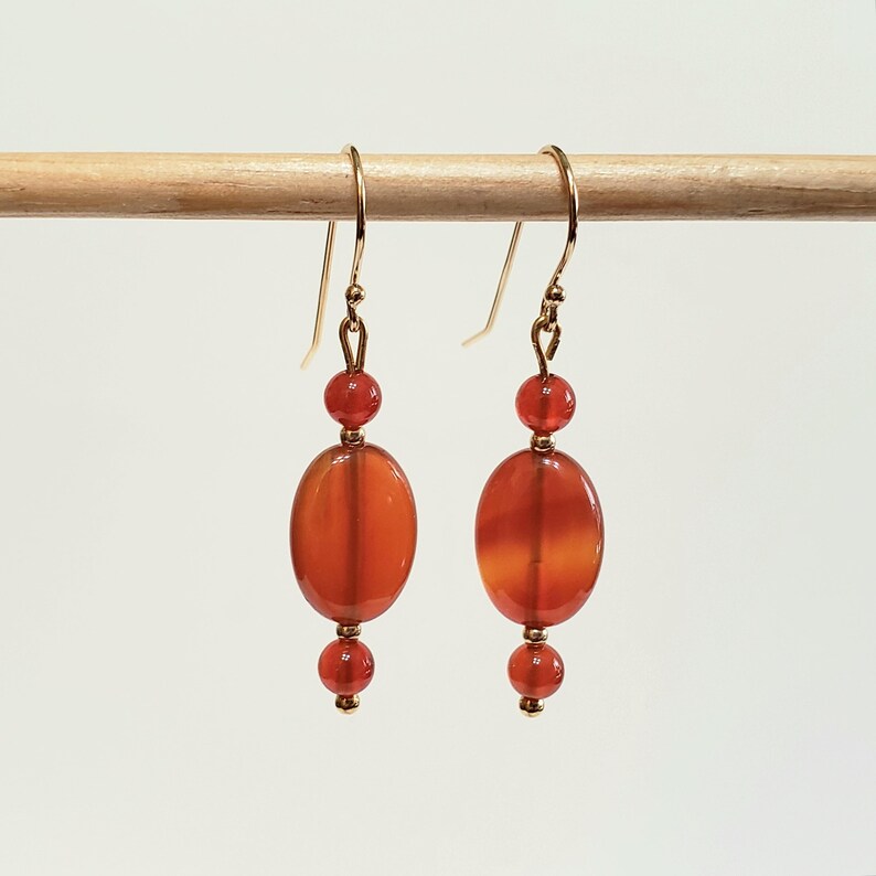 May include: A pair of dangle earrings featuring orange-red carnelian beads. Each earring consists of a small round bead above a larger oval bead, followed by a smaller round bead at the base. The beads are suspended from gold-toned metal hooks.