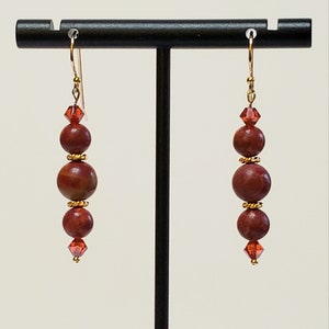 May include: Red and gold beaded earrings with a crystal accent. The earrings are on a black stand.