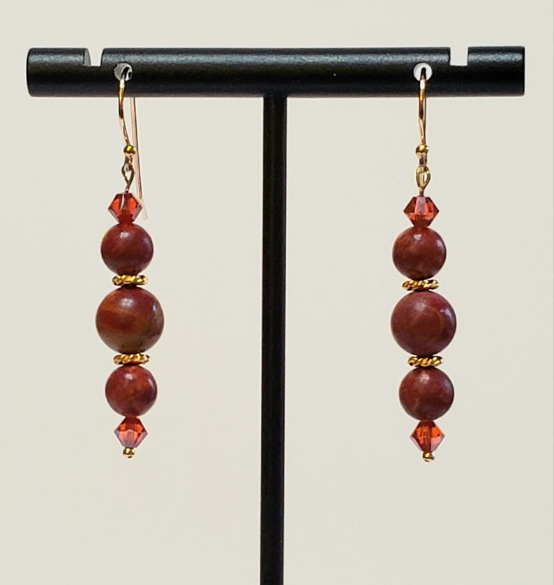 May include: Red and gold beaded earrings with a crystal accent. The earrings are on a black stand.