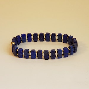 May include: A blue lapis lazuli bracelet with gold beads. The bracelet is made of rectangular lapis lazuli stones and gold beads.
