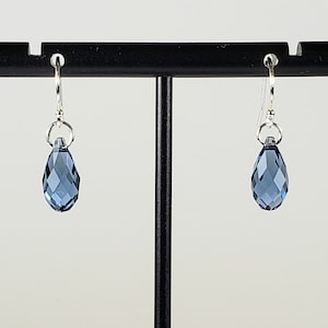 May include: A pair of dangle earrings featuring faceted, teardrop-shaped blue crystals. The crystals are suspended from silver-colored hooks. The earrings are displayed on a black earring stand against a white background. The crystals have a multi-faceted design.