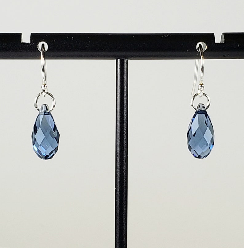 May include: A pair of dangle earrings featuring faceted, teardrop-shaped blue crystals. The crystals are suspended from silver-colored hooks. The earrings are displayed on a black earring stand against a white background. The crystals have a multi-faceted design.