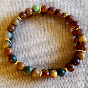 May include: A multi-colored stone bracelet with gold accents. The bracelet is made of round beads with a variety of colors, including red, brown, green, and yellow. The beads are separated by small gold spacers.