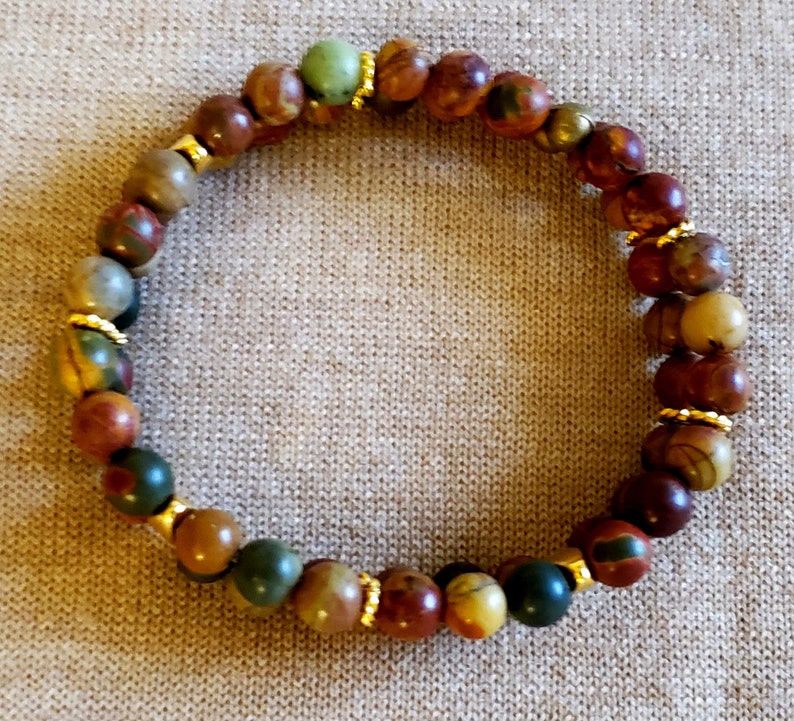 May include: A multi-colored stone bracelet with gold accents. The bracelet is made of round beads with a variety of colors, including red, brown, green, and yellow. The beads are separated by small gold spacers.