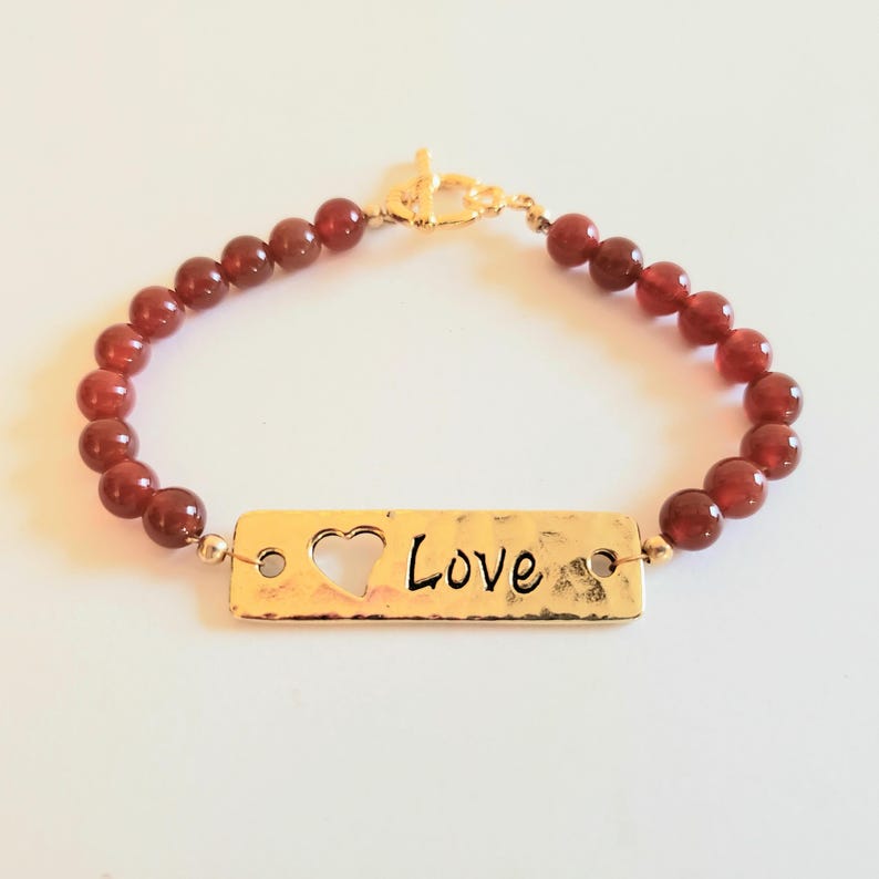 May include: A bracelet featuring a series of round, deep red beads and a central rectangular gold-toned metal plate. The plate has a hammered texture, a cutout heart shape on the left, and the word Love inscribed in black script on the right.