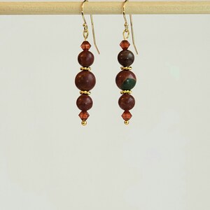 May include: A pair of gold dangle earrings with three brown and green jasper beads and a small crystal bead at the bottom of each earring.