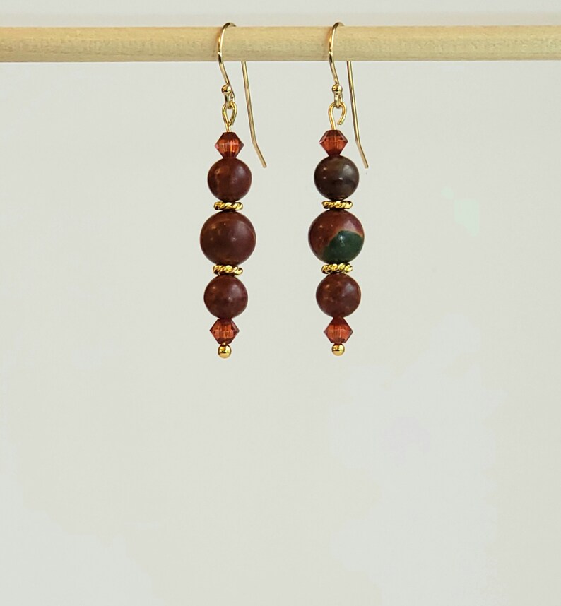 May include: A pair of gold dangle earrings with three brown and green jasper beads and a small crystal bead at the bottom of each earring.