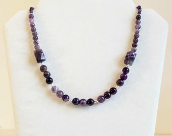 May include: A beaded necklace featuring round and rectangular amethyst beads in various shades of purple, ranging from light lavender to deep violet. The beads are strung together with small silver-toned spacers, creating a symmetrical pattern.
