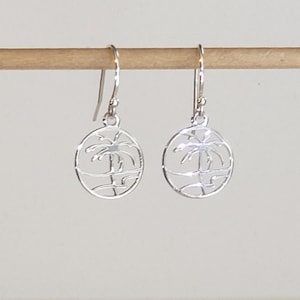 May include: A pair of silver earrings with a cutout design of a palm tree on a beach. The earrings are hanging from a wooden surface.