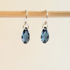 May include: A pair of teardrop-shaped earrings with faceted, dark blue crystals. The earrings hang from silver-colored ear wires. The crystals are suspended from small silver rings.