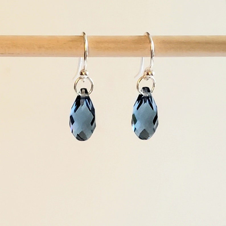 May include: A pair of teardrop-shaped earrings with faceted, dark blue crystals. The earrings hang from silver-colored ear wires. The crystals are suspended from small silver rings.