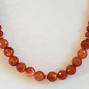 May include: A necklace featuring a strand of round, faceted, burnt orange carnelian beads. The beads are arranged in a uniform sequence and are accented by small, polished gold-toned metal spacers placed at intervals along the length of the strand.