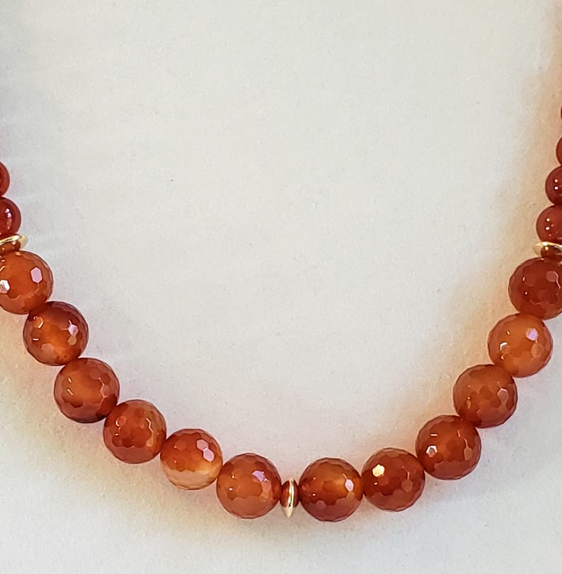May include: A necklace featuring a strand of round, faceted, burnt orange carnelian beads. The beads are arranged in a uniform sequence and are accented by small, polished gold-toned metal spacers placed at intervals along the length of the strand.