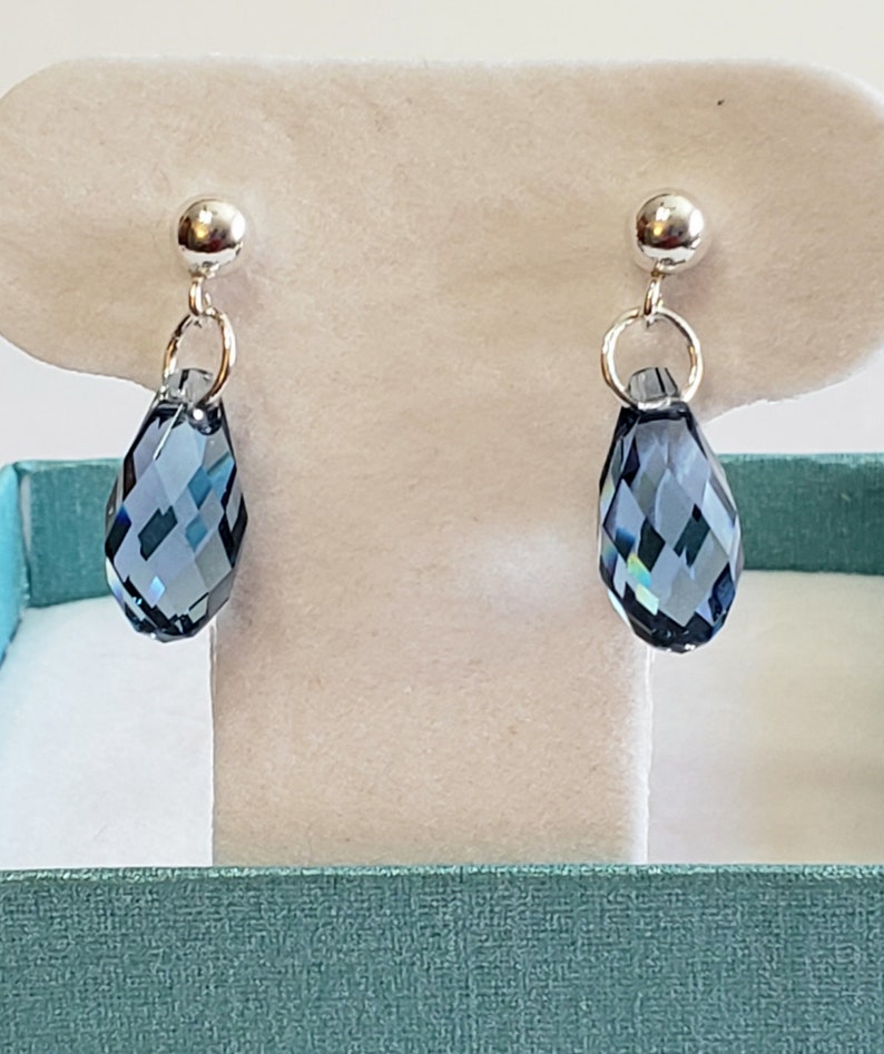 May include: A pair of elegant drop earrings. Each earring features a faceted, teardrop-shaped, dark blue crystal pendant suspended from a silver-colored post. The earrings are displayed on a white background.
