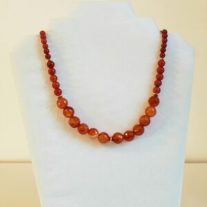 May include: A necklace featuring a strand of round, faceted, deep orange carnelian beads that increase in size toward the center. The beads are separated by small, thin, gold-toned metal spacers, creating a graduated design with a polished finish.