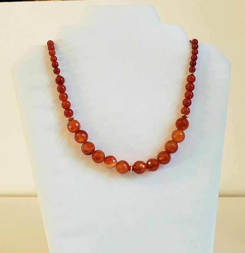 May include: A necklace featuring a strand of round, faceted, deep orange carnelian beads that increase in size toward the center. The beads are separated by small, thin, gold-toned metal spacers, creating a graduated design with a polished finish.