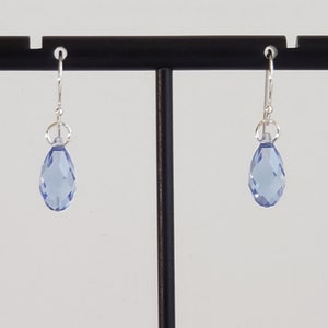 May include: A pair of light blue teardrop earrings with faceted surfaces. The earrings hang from silver-colored hooks and are displayed on a black stand against a white background. The earrings are a simple, elegant design.