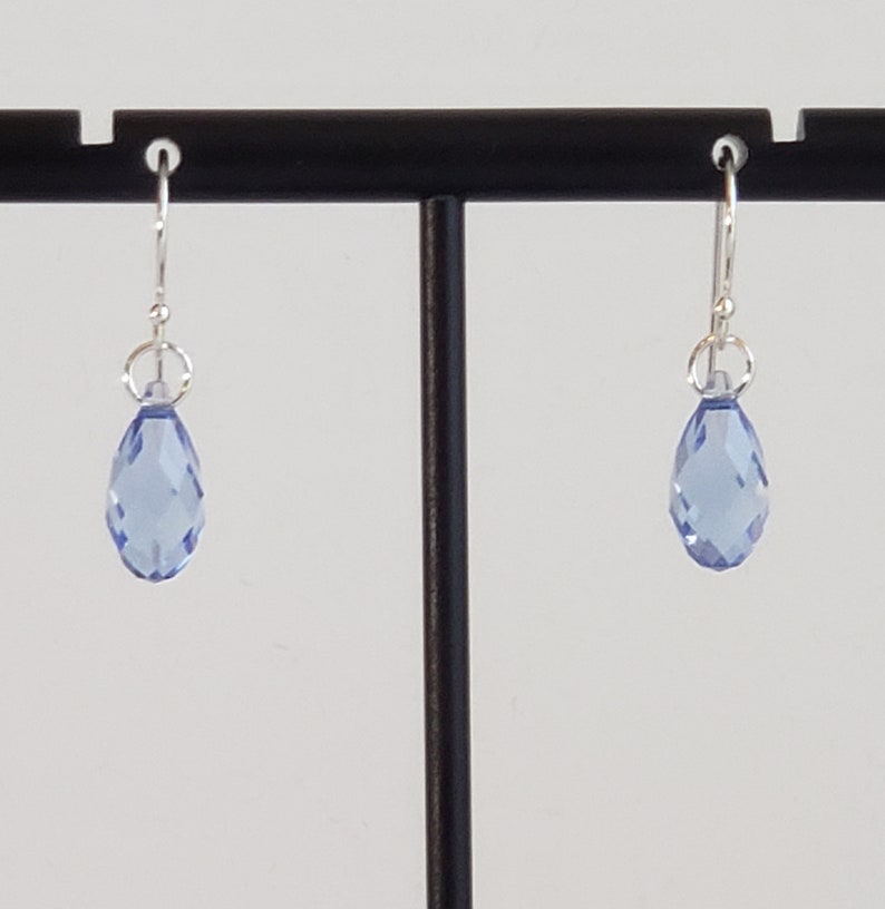 May include: A pair of light blue teardrop earrings with faceted surfaces. The earrings hang from silver-colored hooks and are displayed on a black stand against a white background. The earrings are a simple, elegant design.