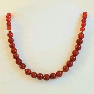 May include: A necklace composed of smooth, round, deep orange-red beads arranged in a single strand. The beads are uniform in size and shape, interspersed with small, polished gold-toned metal spacer beads at regular intervals along the length.