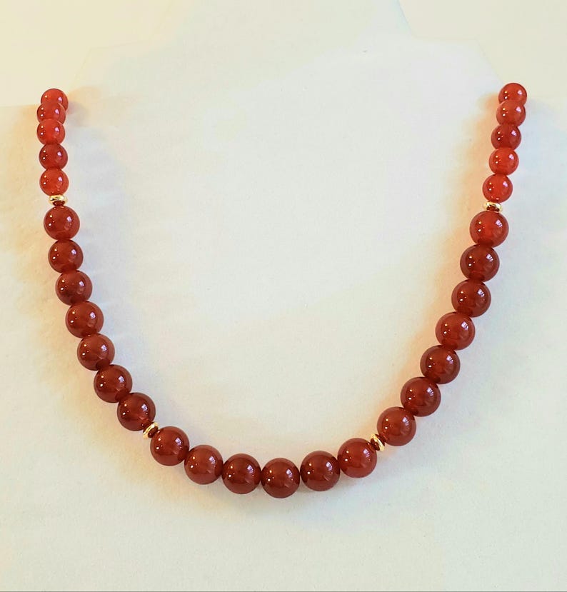 May include: A necklace composed of smooth, round, deep orange-red beads arranged in a single strand. The beads are uniform in size and shape, interspersed with small, polished gold-toned metal spacer beads at regular intervals along the length.