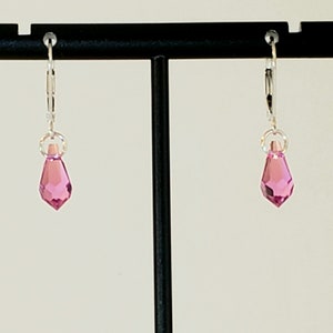 May include: A pair of silver hoop earrings with a pink crystal dangle. The earrings are displayed on a black stand.