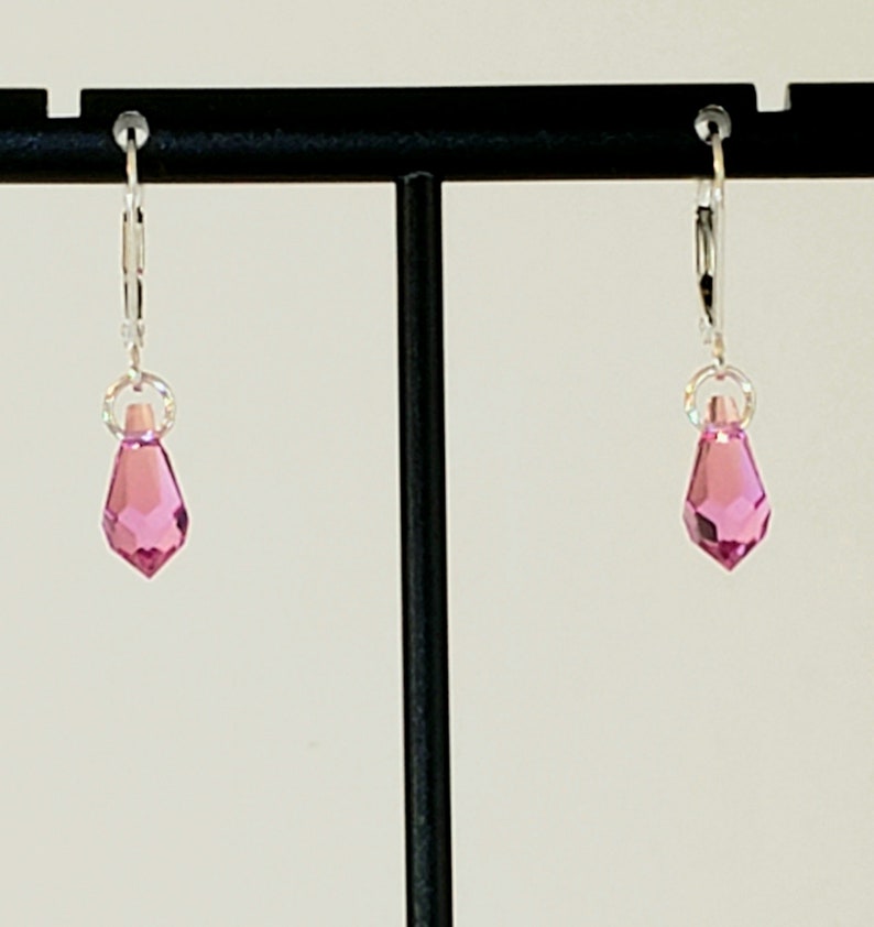 May include: A pair of silver hoop earrings with a pink crystal dangle. The earrings are displayed on a black stand.